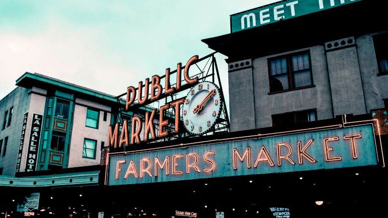 The image displays a street view of a "PUBLIC MARKET" sign with an adjacent "FARMERS MARKET" neon sign and surrounding buildings in an urban area.