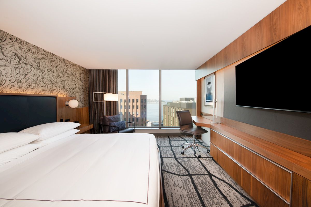 A modern hotel room with a large bed, desk, chair, wall-mounted TV, and window with a city and water view.