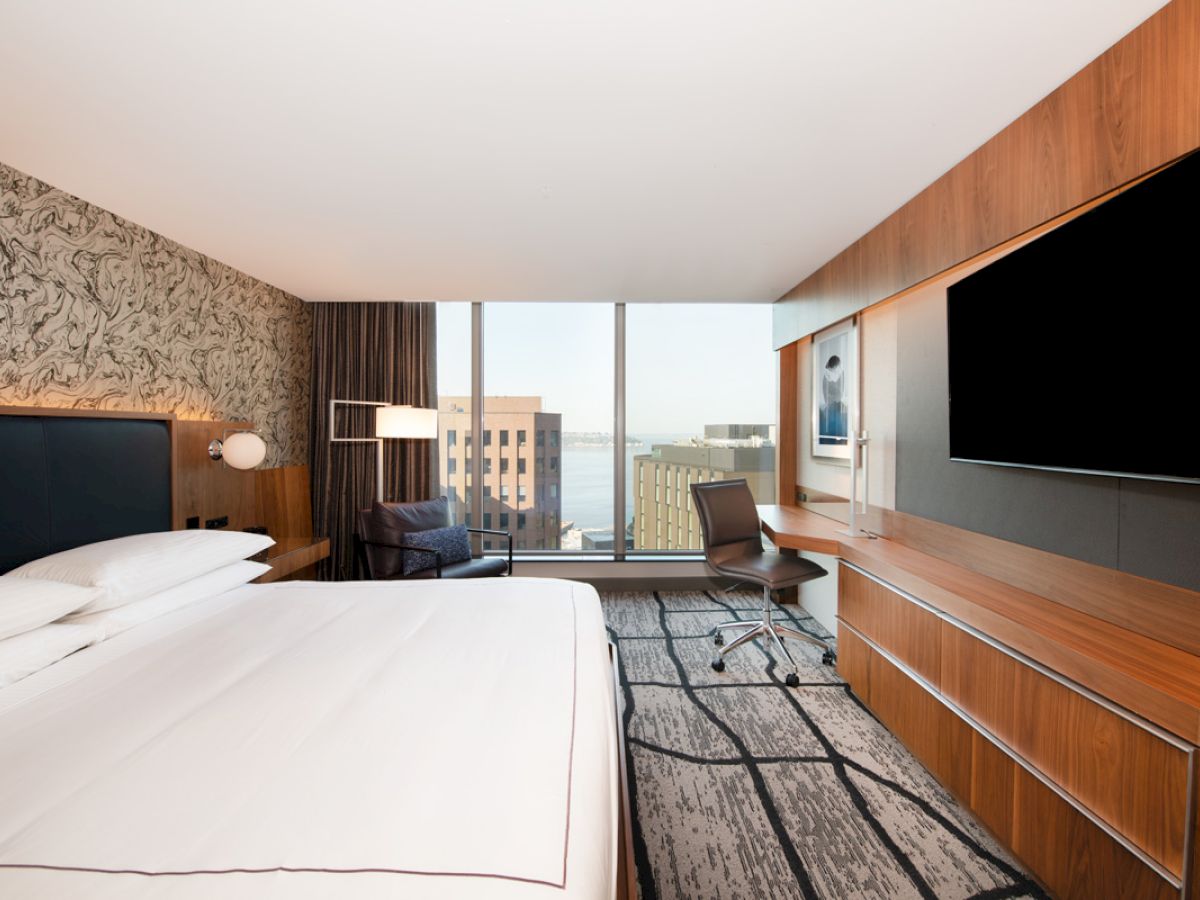 A modern hotel room with a large bed, desk, chair, wall-mounted TV, and window with a city and water view.