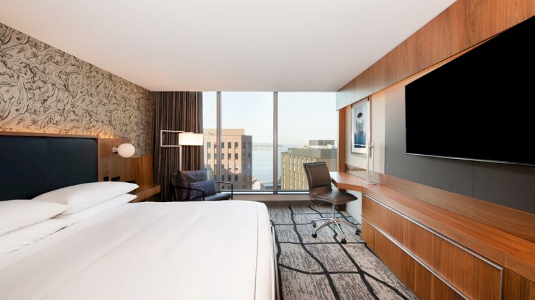 A modern hotel room with a large bed, desk, chair, wall-mounted TV, and window with a city and water view.