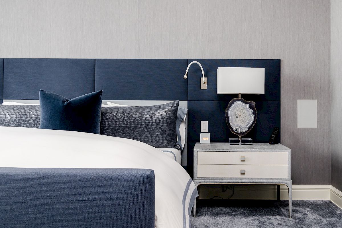 A modern bedroom featuring a blue and white color scheme, with a bed, nightstand, lamp, and decorative pillows.