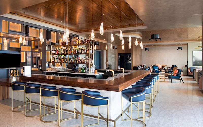 A modern bar with a wooden counter, blue stools, a wide variety of bottles, pendant lights, and a lounge area with seating in the background.