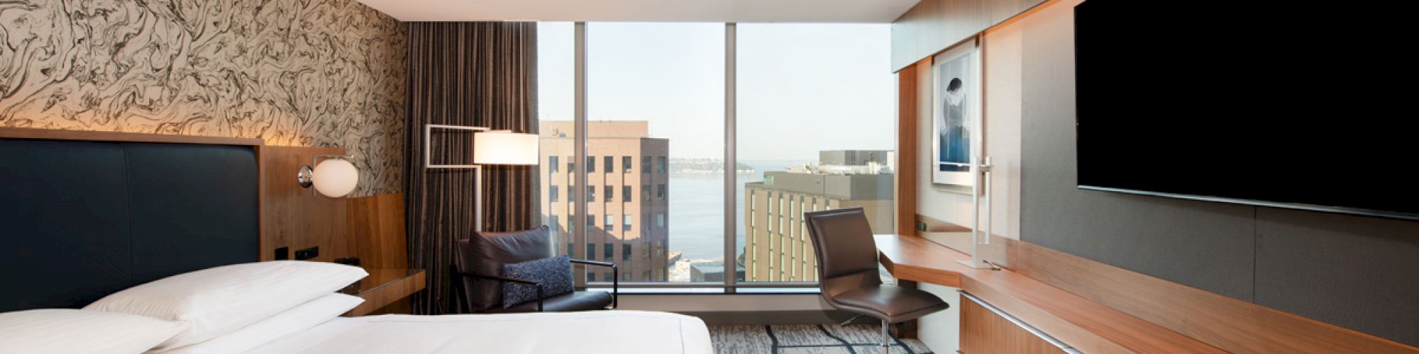 A modern hotel room with a large bed, desk, chair, flat-screen TV, and a window displaying a city view with buildings and some water in the distance.