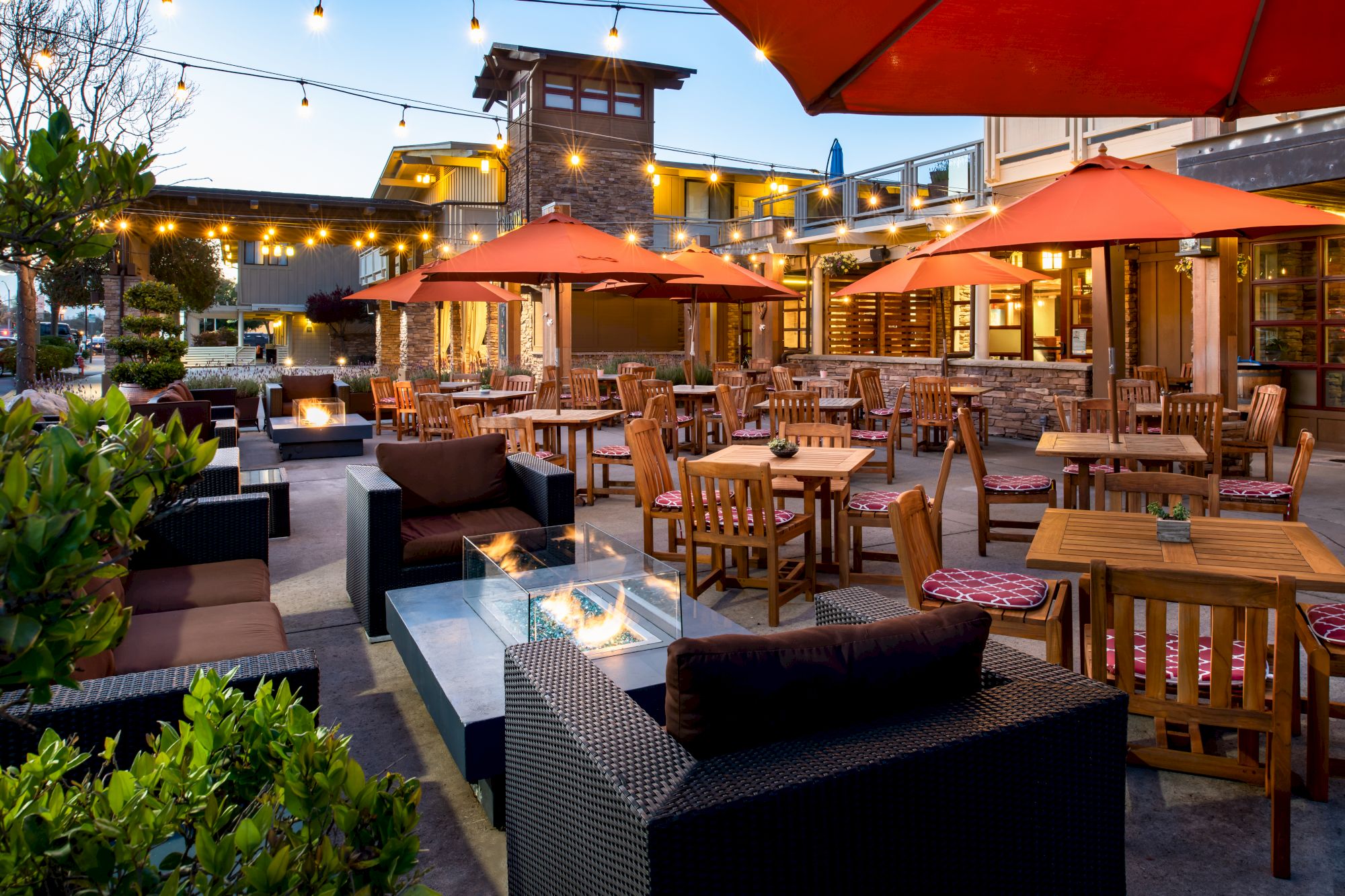 An outdoor dining area with wooden tables and chairs, orange umbrellas, string lights, and a fire pit, all set in a cozy and inviting atmosphere.