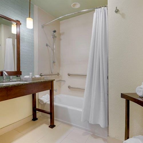 The image shows a modern bathroom with a shower-tub combination, a vanity with a large mirror, and a small wooden table with neatly rolled towels.