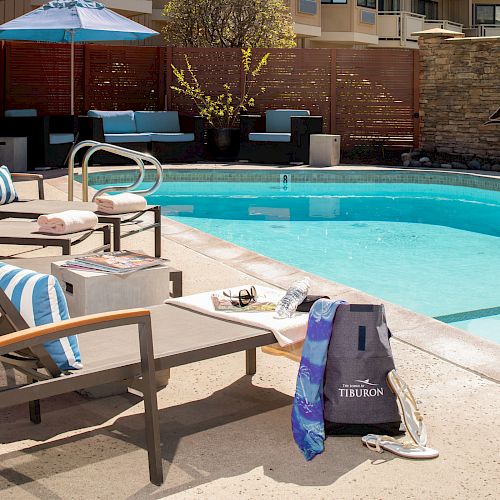 A poolside scene with lounge chairs, towels, a bag labeled "TIBURON," and a book on a table. There is a blue umbrella and a stone wall with a sailboat.