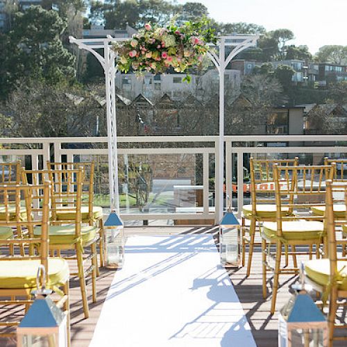 An outdoor wedding setup with a white aisle, wooden chairs, flower arch, and lanterns, surrounded by scenic greenery and houses in the background.