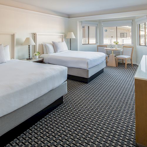 The image shows a hotel room with two beds, a TV, a small table with chairs near the window, and modern decor featuring a patterned carpet.