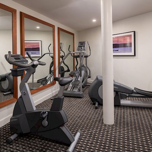 A small gym with exercise bikes, elliptical machines, large mirrors, and a framed picture on the wall. The room has carpeted flooring.