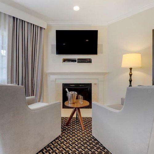 This image shows a cozy room with two grey armchairs facing a small table, a fireplace, a mounted TV, and a lamp near a door.