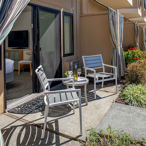 An outdoor seating area with two chairs and a small table, a bottle, and a glass, near a sliding door leading to an indoor room with a TV.