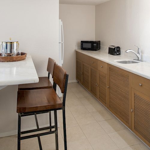 A kitchenette with a countertop, two bar stools, a fridge, microwave, toaster, sink, and cabinets with wooden doors can be seen in the image.