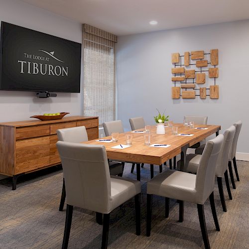 A modern conference room with a wooden table, eight chairs, a wall-mounted screen displaying 