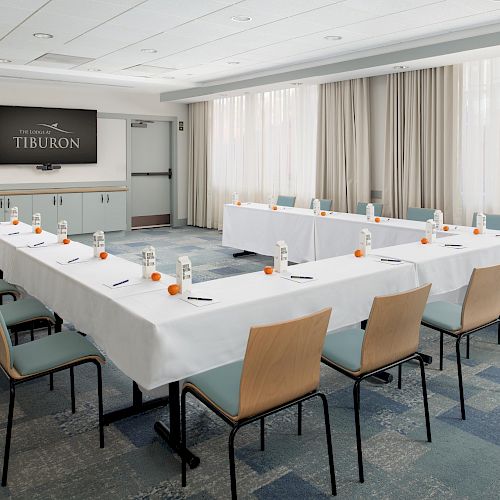 A conference room with U-shaped setup, featuring tables with white tablecloths, chairs, bottled water, oranges, and notepads.