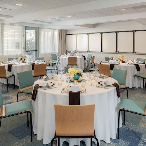 The image shows a well-lit conference or banquet room with round tables set for dining, each decorated with flowers, surrounded by chairs.