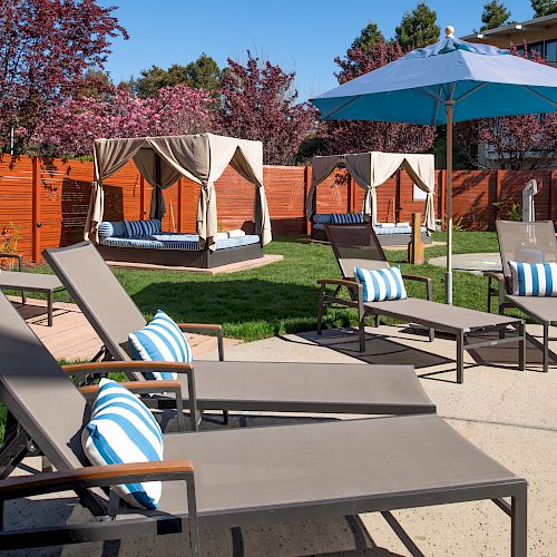 The image depicts an outdoor pool area with lounge chairs, striped cushions, cabanas, and a large umbrella, surrounded by greenery and a wooden fence.