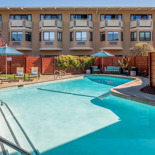 The image features a swimming pool with lounge chairs and umbrellas, surrounded by a modern building with multiple balconies and a privacy fence.