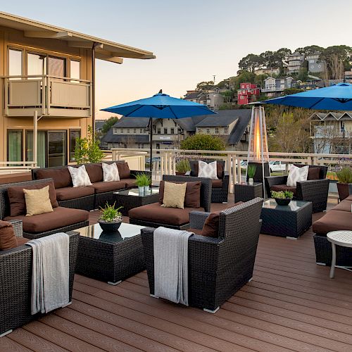 The image shows an outdoor patio with wicker furniture, blue umbrellas, and gas heaters, set on a wooden deck with a scenic background of greenery and buildings.