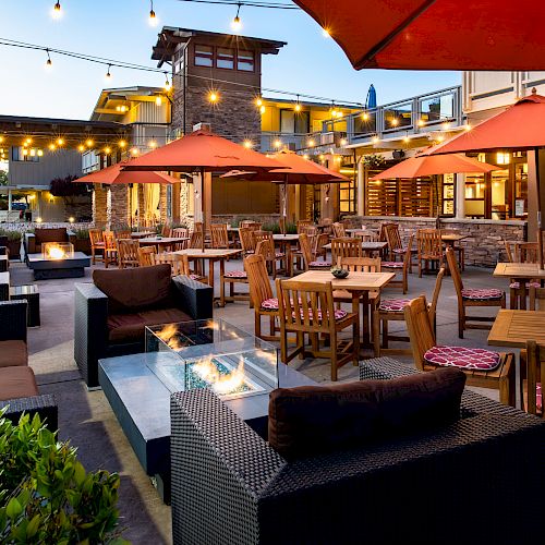 The image shows an outdoor dining area with wooden furniture, red umbrellas, string lights, potted plants, and a modern building in the background.