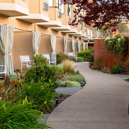 A curved walkway leads past chairs outside private patio areas with curtains in a landscaped garden featuring various plants and flowers.