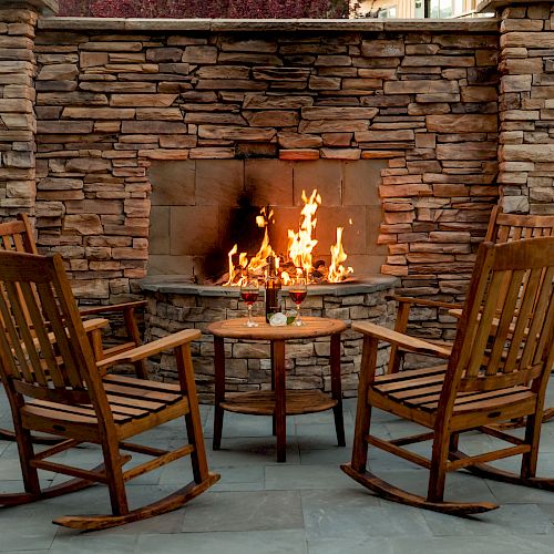 Two wooden rocking chairs face a stone fireplace with a small table and drinks between them on a patio.