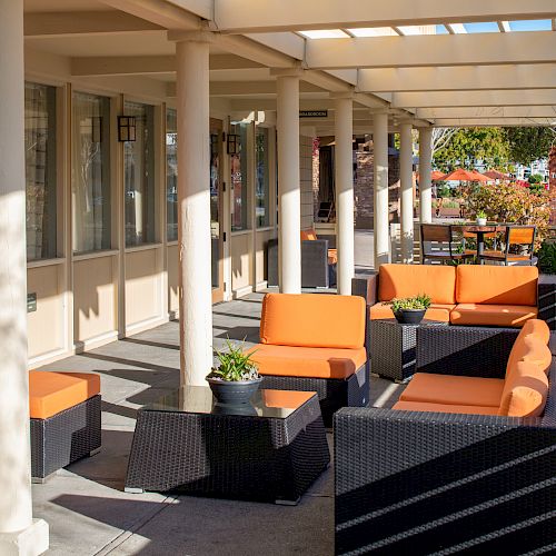 Outdoor patio with wicker furniture, bright orange cushions, a few potted plants on tables, under a pergola with sunlight and shadows.