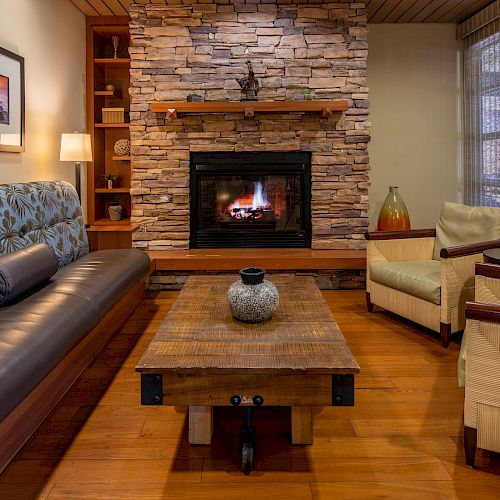 A cozy living room with a stone fireplace, wooden coffee table, sofa, armchairs, lamps, and wall art, creating a warm and inviting atmosphere.