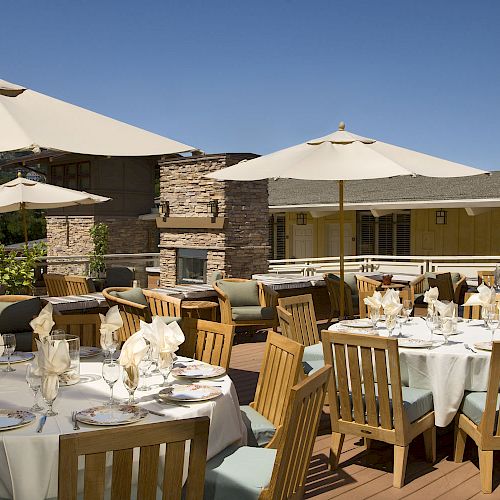 An outdoor patio with wooden tables and chairs set up for dining, featuring white tablecloths, napkins, and large umbrellas under a clear blue sky.