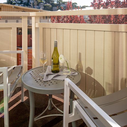 A cozy balcony setup with two white chairs and a small round table featuring a bottle of wine, two glasses, and a napkin under a sunny backdrop.