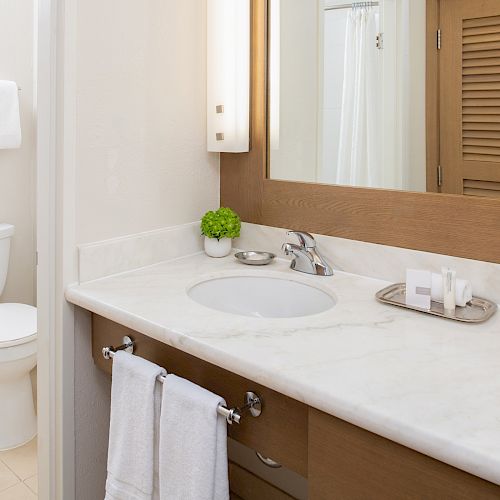 The image shows a clean, modern bathroom with a white toilet, a sink with a marble countertop, a mirror, and towels neatly arranged.