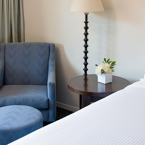 A cozy hotel room corner with a blue armchair, matching ottoman, side table with a flower, a lamp, and part of a neatly made bed.