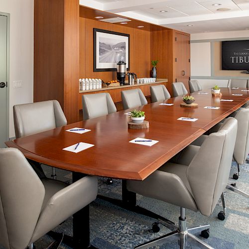 The image shows a conference room with a long wooden table, surrounded by grey chairs, and a screen displaying 