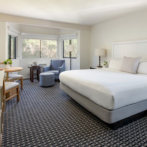 The image shows a neatly arranged hotel room with a king-sized bed, a TV, a table and chairs, and a sitting area by the window.