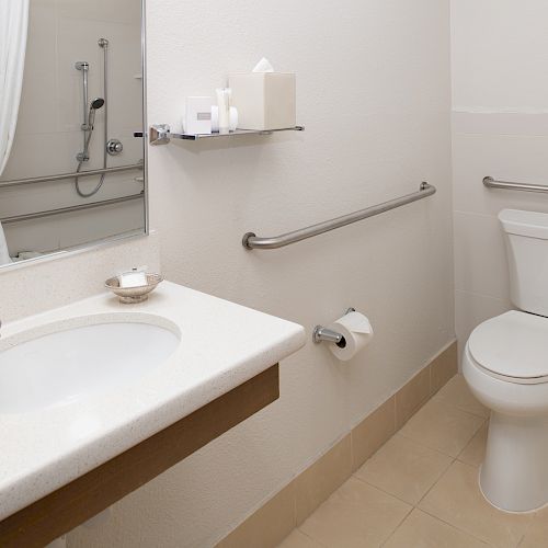 A modern bathroom with a sink, mirror, toilet, and shower area, featuring white fixtures and grab bars for accessibility, ending the sentence.