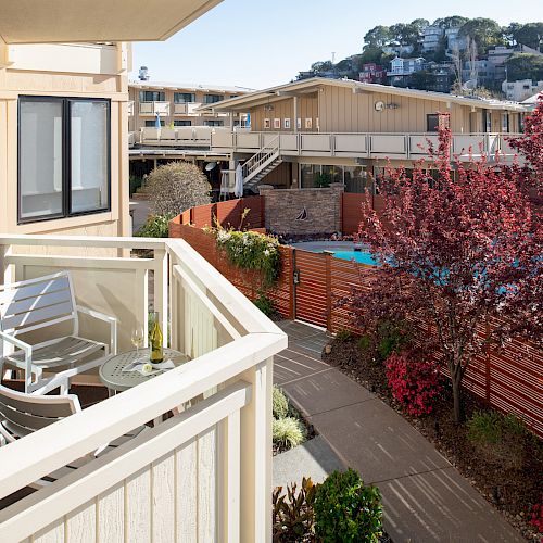 This image shows a residential balcony with two chairs and a table overlooking a courtyard with plants, a pathway, and buildings in the background.