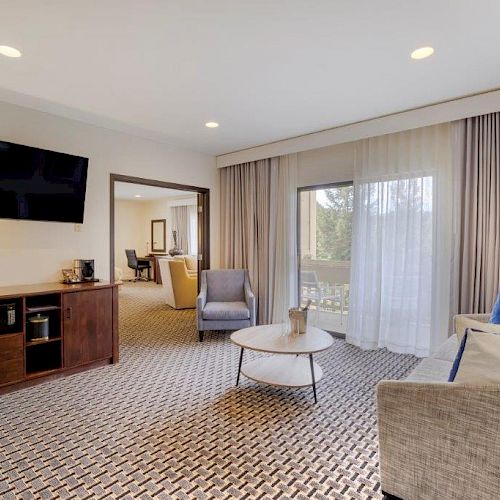 A modern living room with a wall-mounted TV, sofa, chairs, coffee table, patterned rug, floor lamp, and large window with curtains is shown.