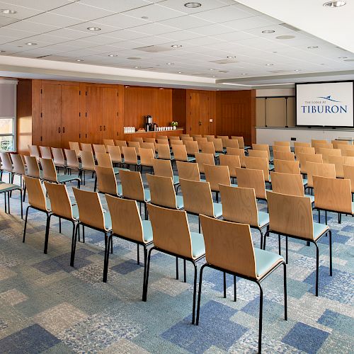 The image shows a conference room set up with rows of chairs facing a screen displaying "TIBURON," with a beverage station in the back.